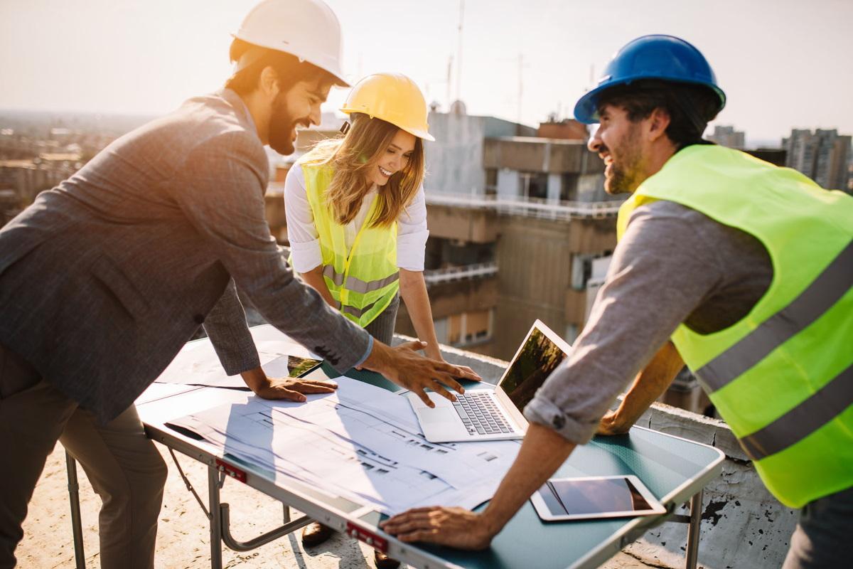 Construction project management team wearing safety helmets and using a laptop to coordinate site plans and estimates.