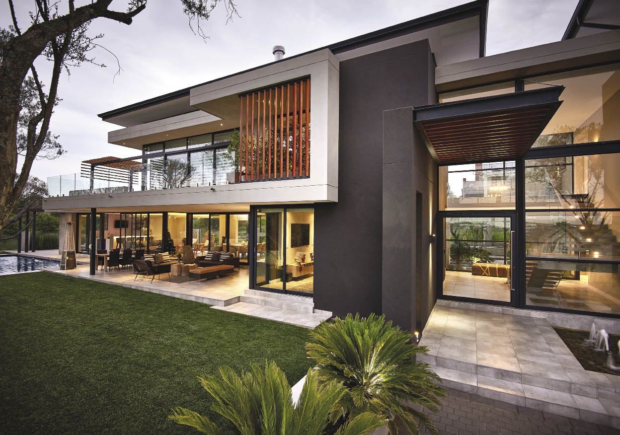 A stunning modern luxury two-story home showcasing seamless interior and exterior designing, featuring floor-to-ceiling glass windows, an illuminated outdoor patio with seating, and a manicured green lawn. To bring these high-end architectural visions to life while staying strictly on budget, builders rely on highly accurate cost estimating services for contractors.
